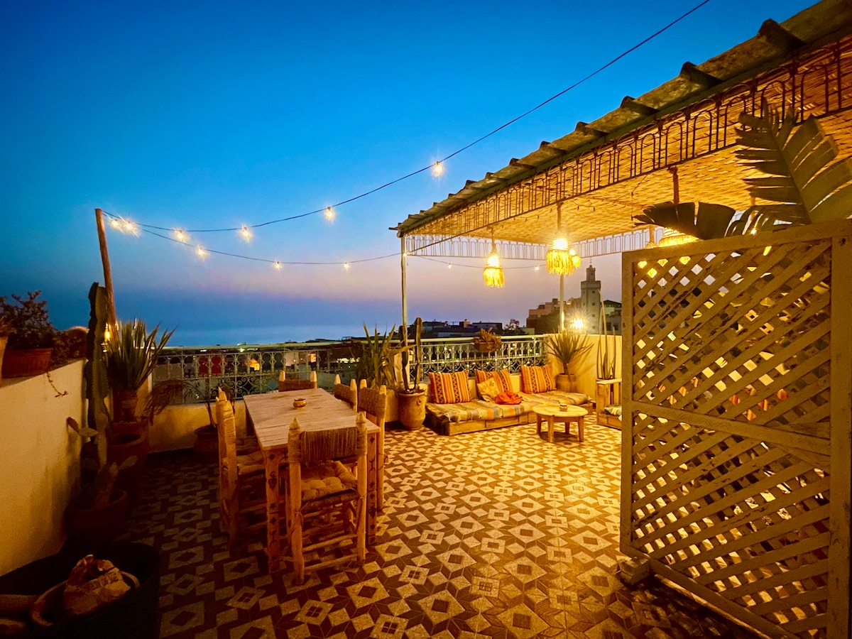An open rooftop terrace features a large wooden table surrounded by comfortable seating. Soft lighting from hanging lamps illuminates the area, complemented by potted plants. The calming evening sky and panoramic views of the ocean and village create a serene backdrop.