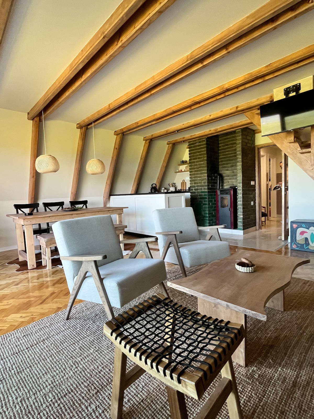 A stylish living area features high wooden beams that create a spacious feel. Two light blue chairs and a natural wood coffee table are arranged on a textured rug. A dining table with black chairs is visible in the background, along with a modern kitchen setup.