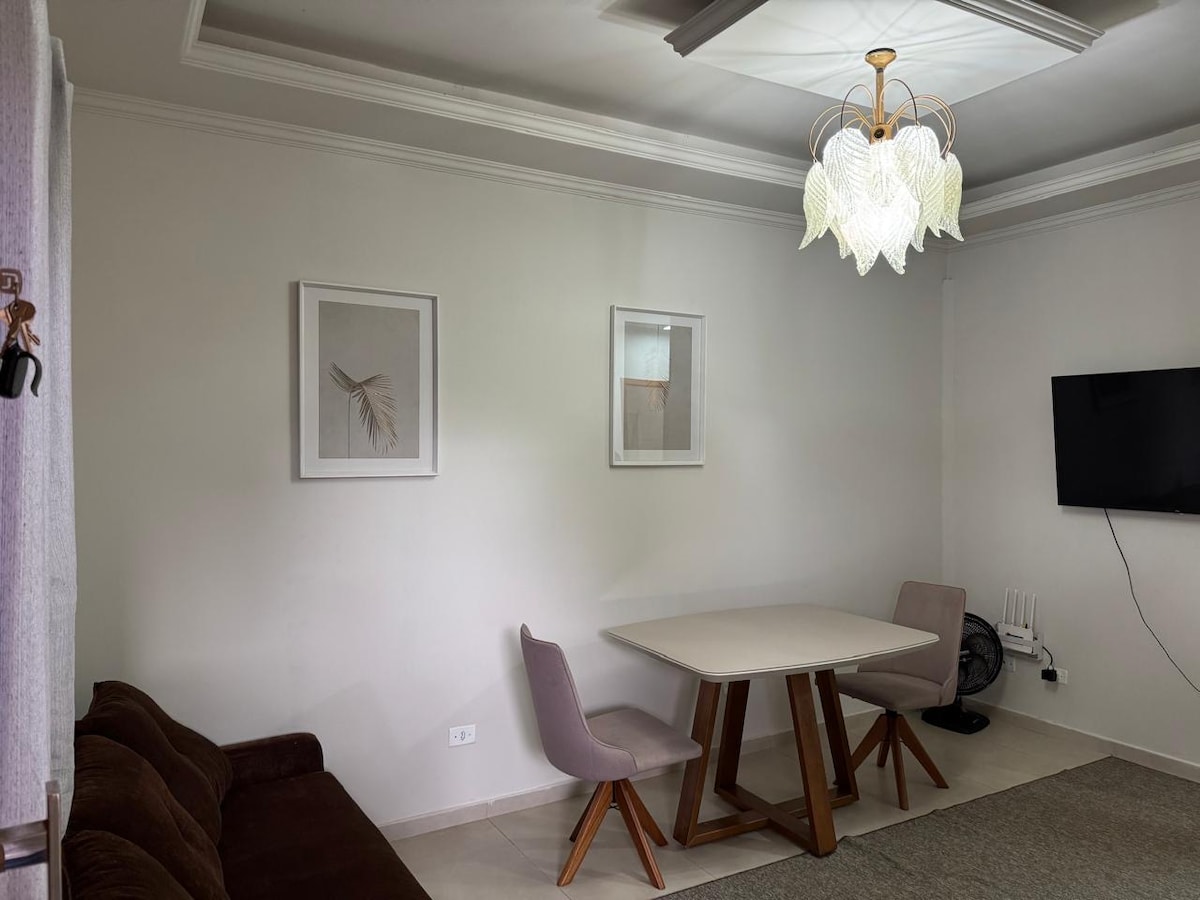 A light-filled living area features a modern dining table surrounded by two upholstered chairs. A comfortable brown sofa is positioned nearby, while two framed art pieces and a stylish chandelier enhance the space. A wall-mounted television is visible on the opposite side.