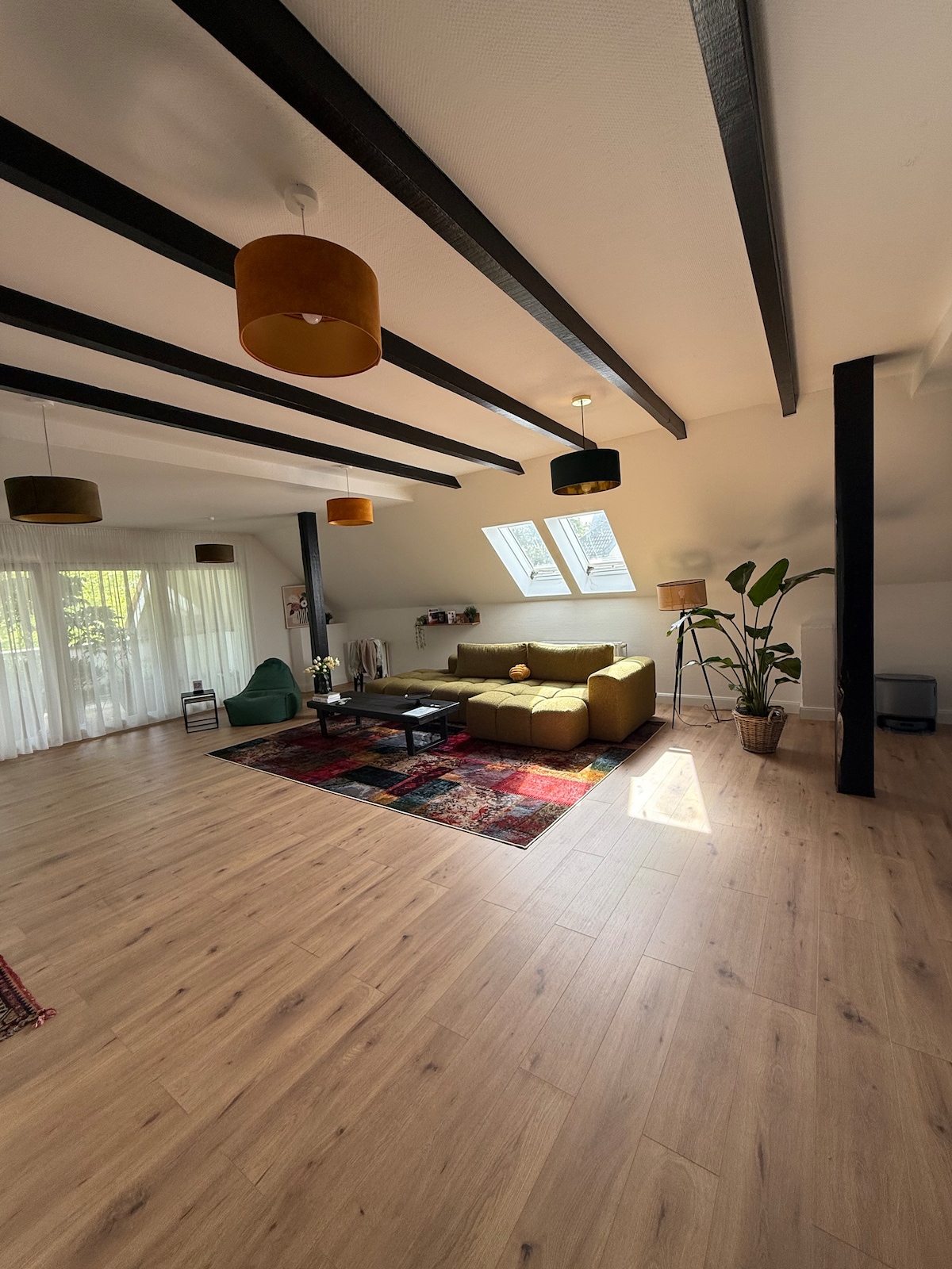 An open living area is shown with wooden flooring and exposed ceiling beams. A green sofa is positioned around a coffee table, alongside a colorful area rug. Natural light fills the space through large windows, and a potted plant adds a touch of greenery.