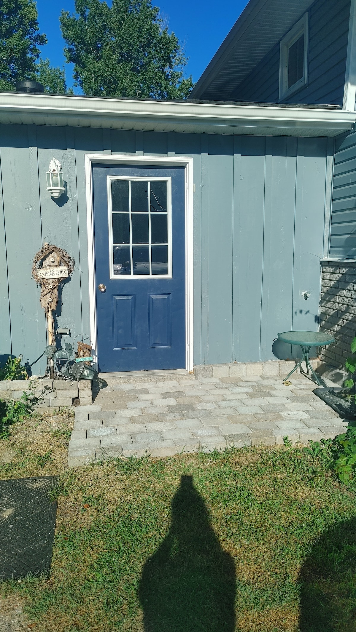 The image features an entrance with a blue door framed by gray siding. A small stone-paved area leads to the door, complemented by a decorative birdhouse and a green wheelbarrow. Lush grass and plants are visible on either side.