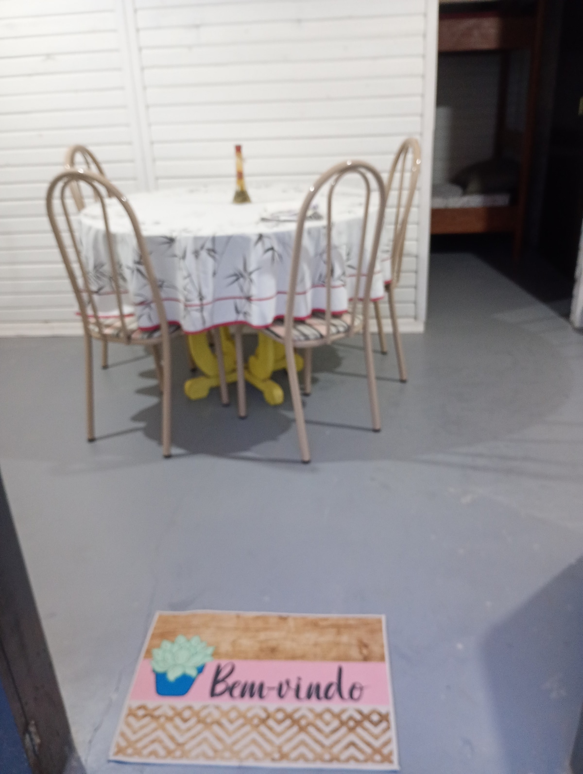 A round dining table is set with a floral tablecloth, accompanied by four metal chairs. A decorative welcome mat is positioned at the entrance, featuring a simple design and welcoming text. The background shows light-colored wooden walls and a glimpse of a sleeping area.