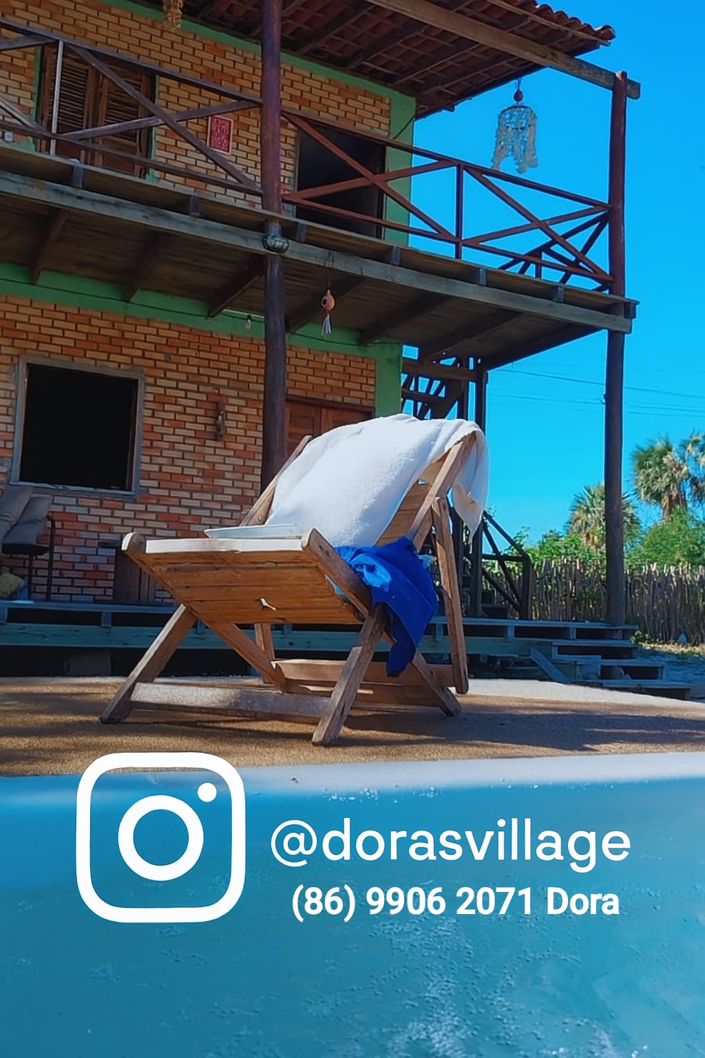 A wooden lounge chair is positioned near the pool, with a blue towel draped over it. The vibrant green and brick structure of the house is visible in the background, featuring a spacious balcony under a clear blue sky.