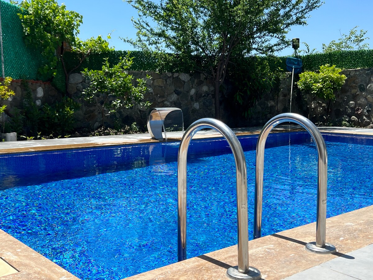 A swimming pool is framed by a stone wall and bordered by lush greenery. The clear, blue water reflects sunlight, while metal handrails provide access. A water feature is visible in the background, adding to the tranquil setting.