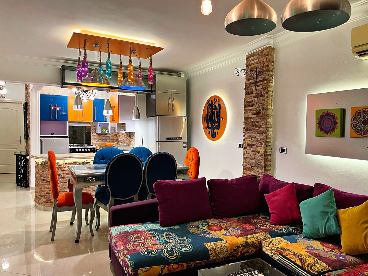 The living area features a large L-shaped sofa adorned with colorful cushions, complemented by a dining table with elegant blue chairs. A well-equipped kitchen is visible, with a distinctive backsplash. Decorative pendant lights hang overhead, providing vibrant visual appeal against the modern decor.