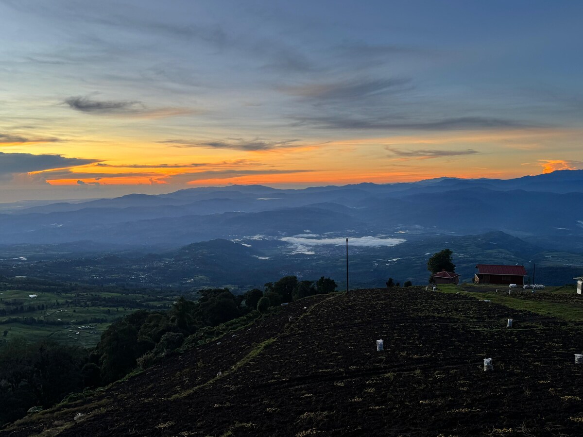 A scenic view captures the landscape at sunset, with layers of mountains shrouded in mist. The sky is painted with soft hues of orange and blue, and a distant structure is visible on the hillside along with patches of greenery.