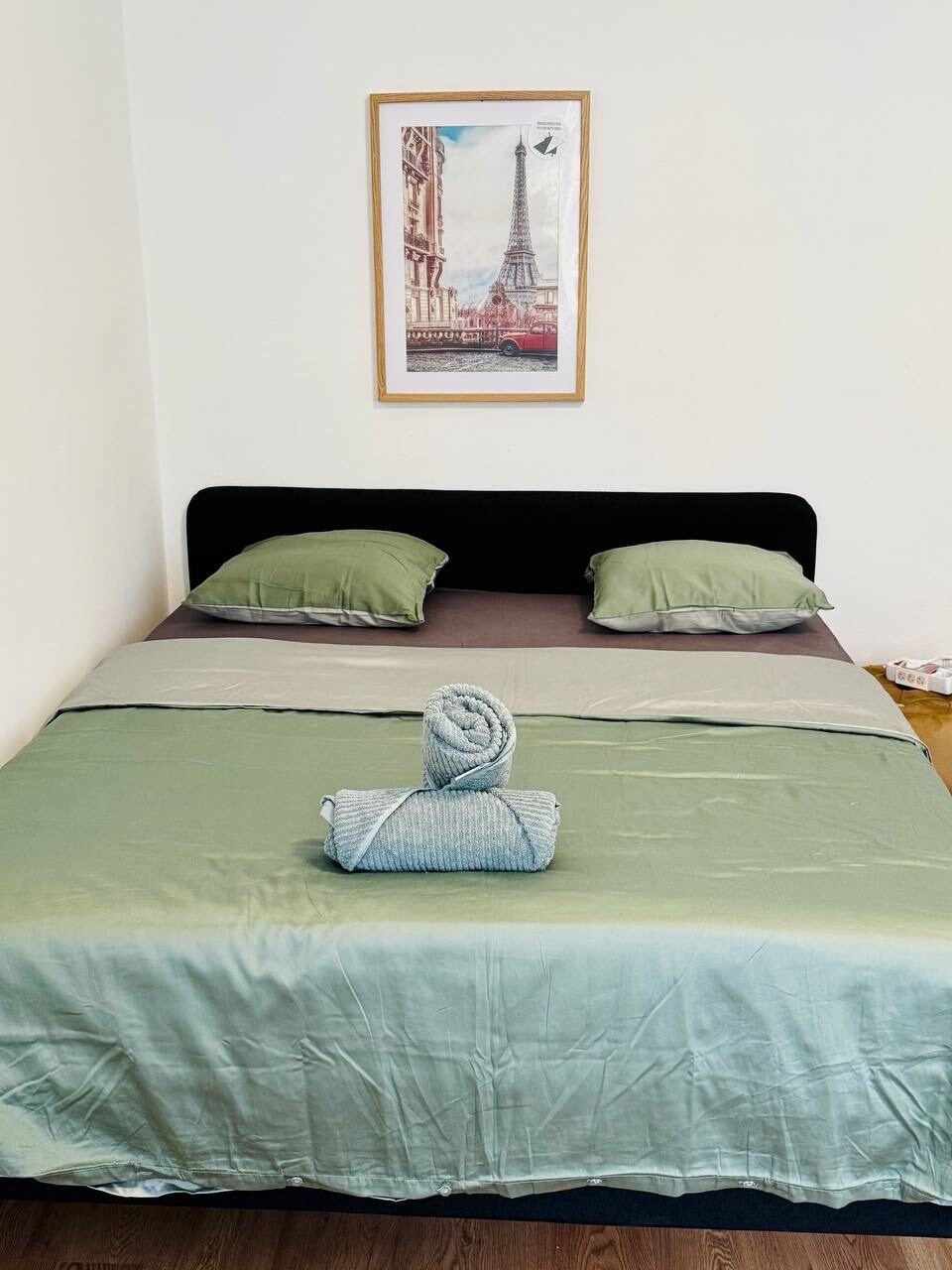 A spacious bed is presented with a light green and grey bedding arrangement. Two matching pillows rest at the head of the bed. A neatly rolled towel is placed at the center. A poster of the Eiffel Tower is displayed prominently on the wall above.