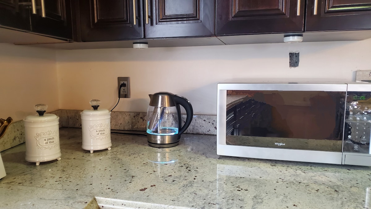 A modern kitchen counter is displayed, featuring a stainless steel kettle and a sleek microwave. Two white canisters for storage sit beside the kettle, all resting on a speckled granite surface. The cabinetry above adds a dark contrast to the light countertop.