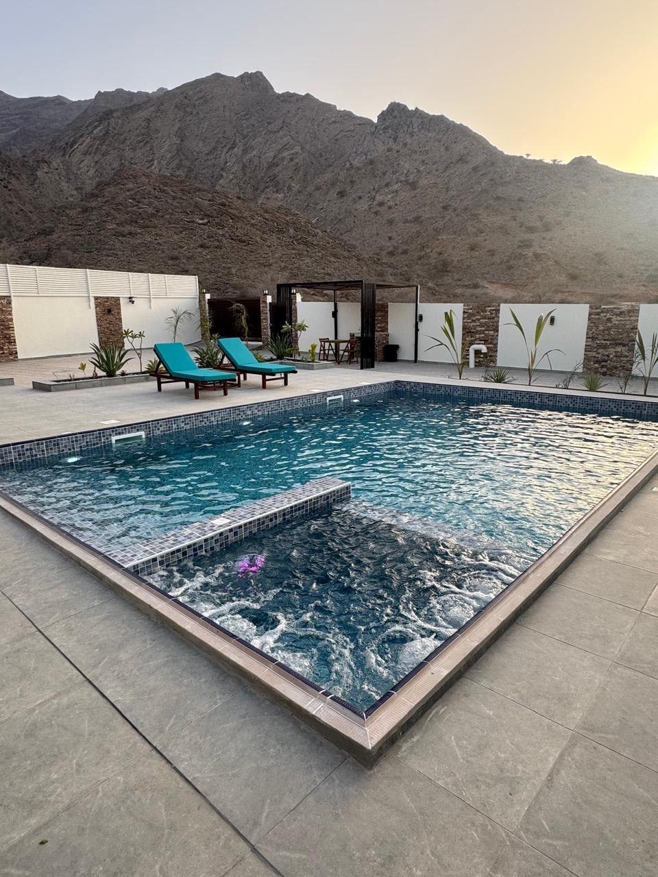 A private pool is framed by elegant stone tiles, shimmering under the soft sunlight. Two comfortable lounge chairs are positioned beside the pool, inviting relaxation. Lush plants are placed around the area, with the majestic mountains rising in the background.