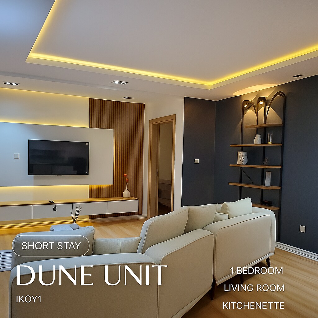 A modern living room features a white sectional sofa paired with a sleek coffee table. Soft lighting highlights a mounted flat-screen TV against a wooden accent wall. An open shelving unit displays decorative items, while the wooden floor contributes to a clean, minimalist aesthetic.