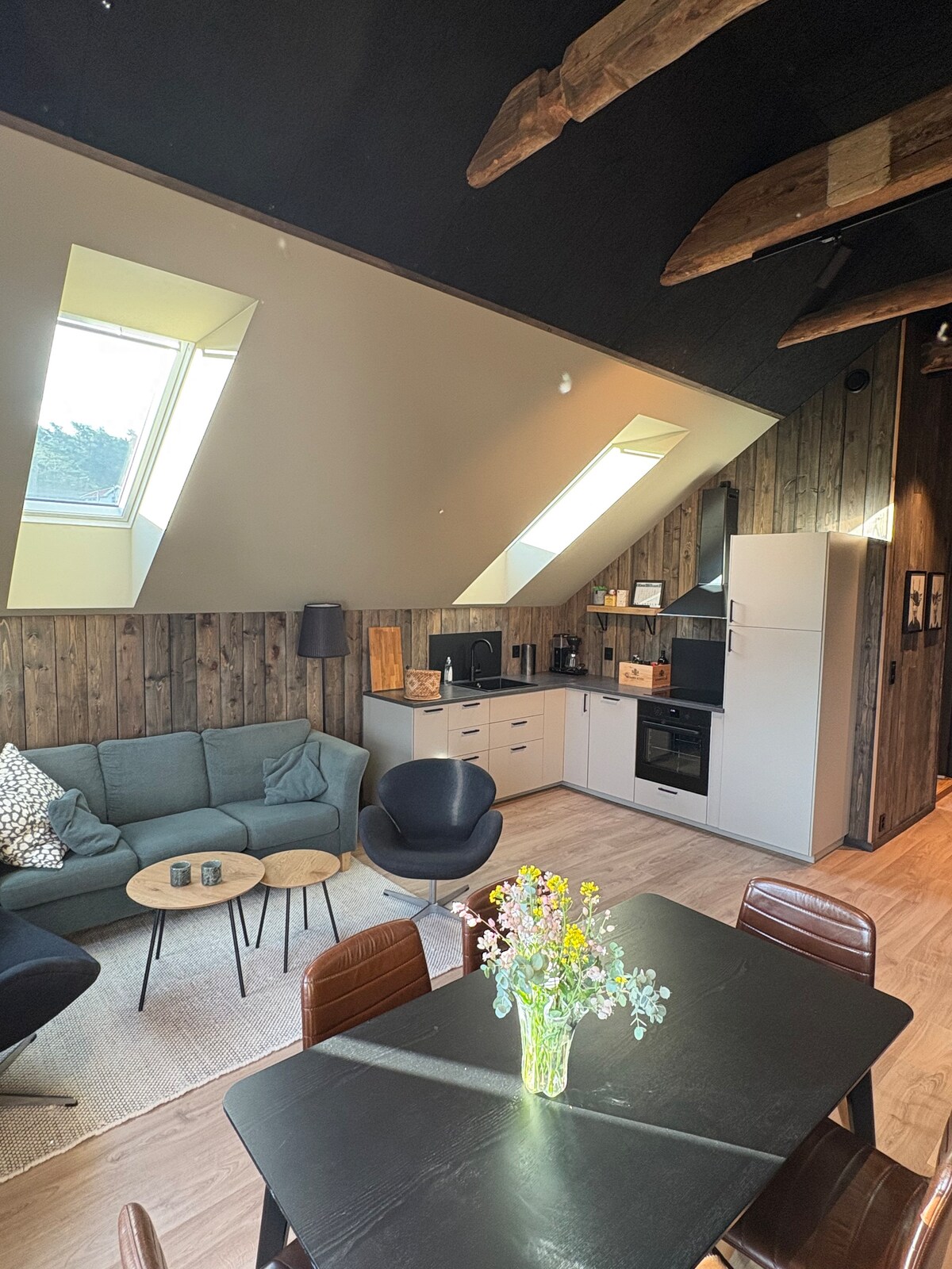 The living area features a cozy seating arrangement with a green sofa and black chairs. A gray rug anchors the space, while natural light enters through two skylights, illuminating the wood-paneled walls. A dining table is visible, adorned with a vase of fresh flowers.