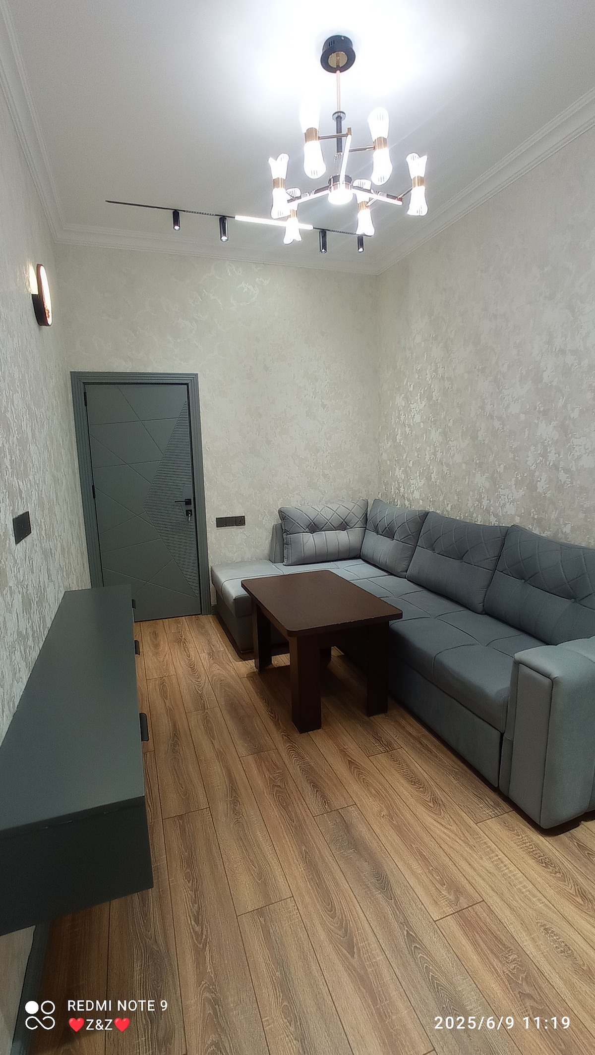 A comfortable living area is presented with a sectional sofa, complemented by a coffee table in the center. A sleek console table is positioned against the wall. Modern lighting fixtures hang from the ceiling, and light wooden flooring enhances the spacious feeling.