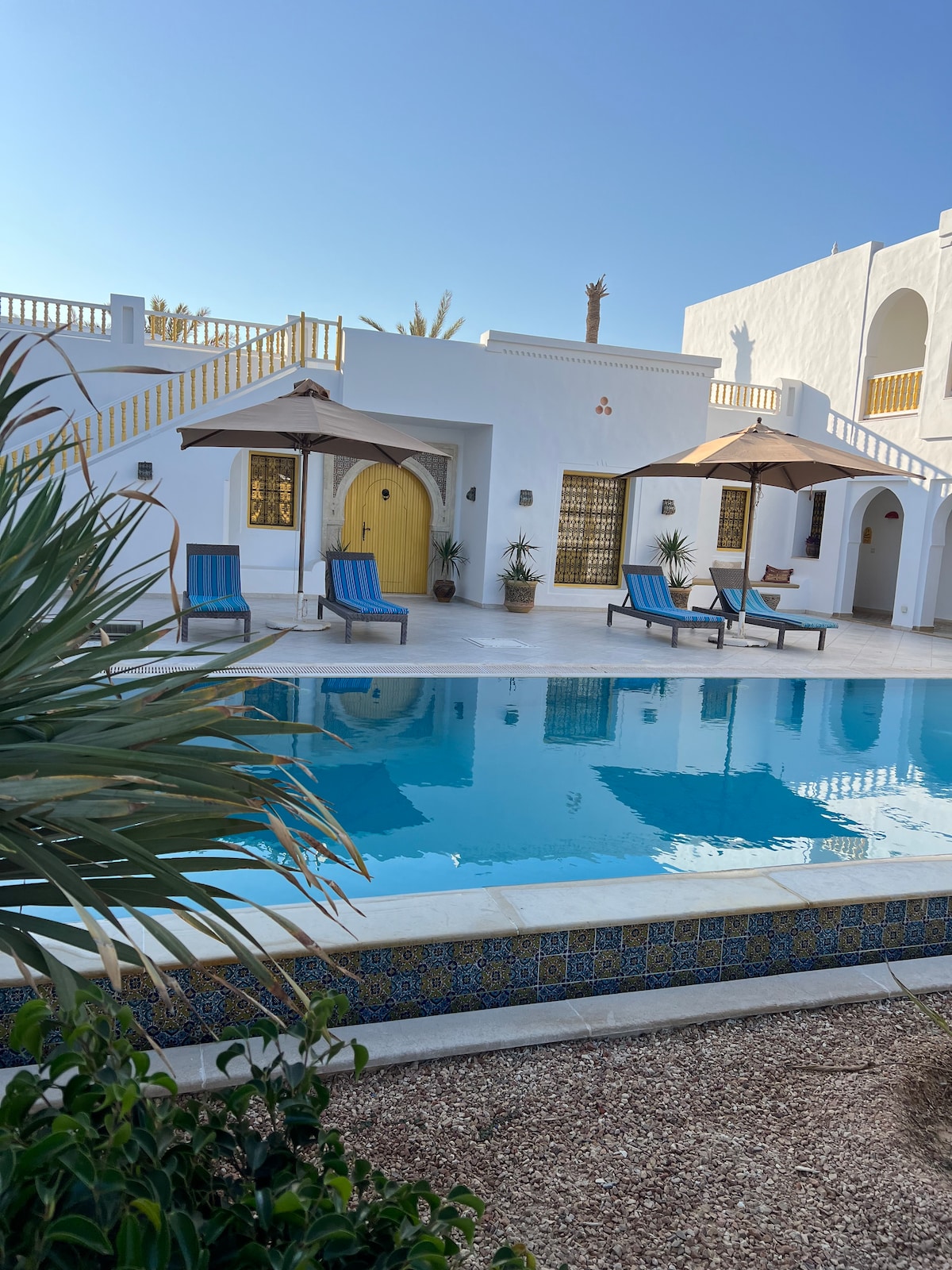 A serene outdoor area is depicted, featuring a crystal-clear swimming pool surrounded by lounge chairs. Two umbrellas provide shade. The building's white walls and a vibrant yellow door create a contrast against the blue sky and lush greenery, enhancing the tranquil atmosphere.
