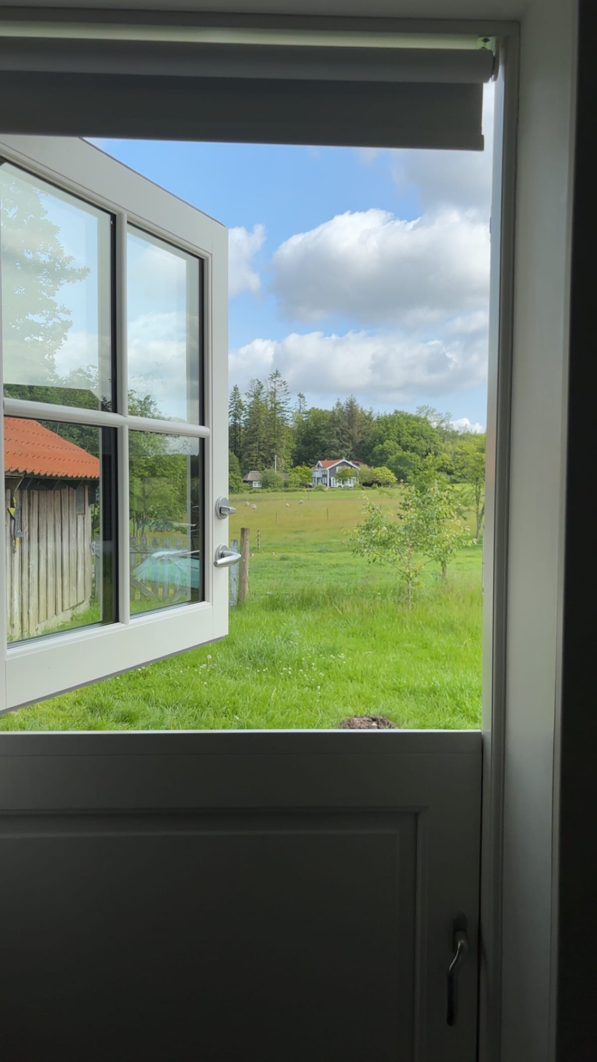 An open window reveals a wide view of lush green fields, with scattered trees and a distant house. The sky is partly cloudy, creating a serene backdrop for the tranquil landscape. The rustic exterior of a neighboring building is visible to the left.