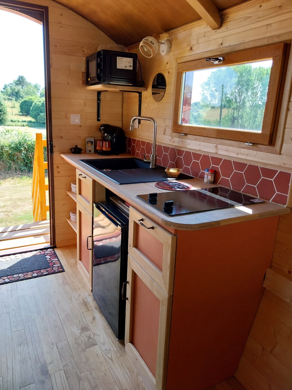 A compact kitchen area features modern appliances including a microwave, coffee maker, and an induction cooktop. Cabinets in warm tones are complemented by a backsplash of hexagonal tiles. A window allows natural light to illuminate the space, offering a view of the surrounding greenery.
