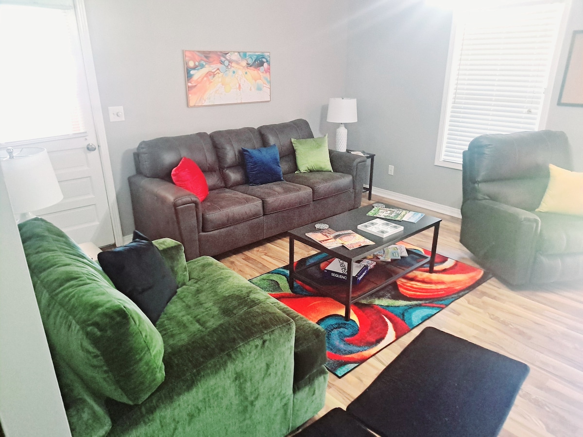 The living room features a dark gray sofa with colorful cushions and two green reclining chairs. A coffee table is situated in the center, adorned with magazines. A vibrant area rug adds color to the light wood flooring, while natural light enters through the window.
