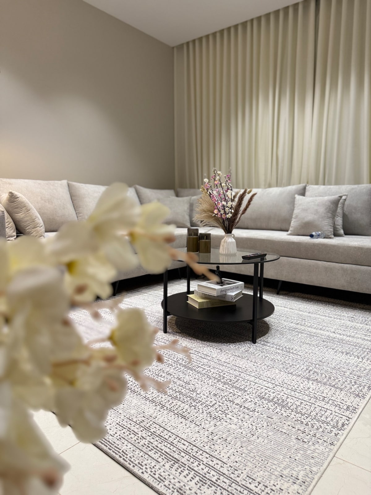 A cozy living area is highlighted by a plush, L-shaped sofa in neutral tones. A low black table is set in front, adorned with a decorative vase and books. Soft, natural light filters through floor-length curtains, adding a sense of warmth to the space.