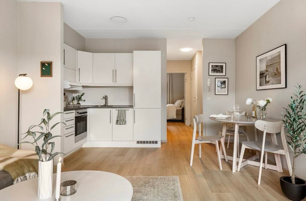 A modern kitchen area features sleek cabinetry and essential appliances, complemented by a wooden dining table set for four. A cozy seating area with a soft throw and decorative accent lighting is visible, contributing to the overall inviting ambiance. Artwork and greenery add subtle touches of decor.