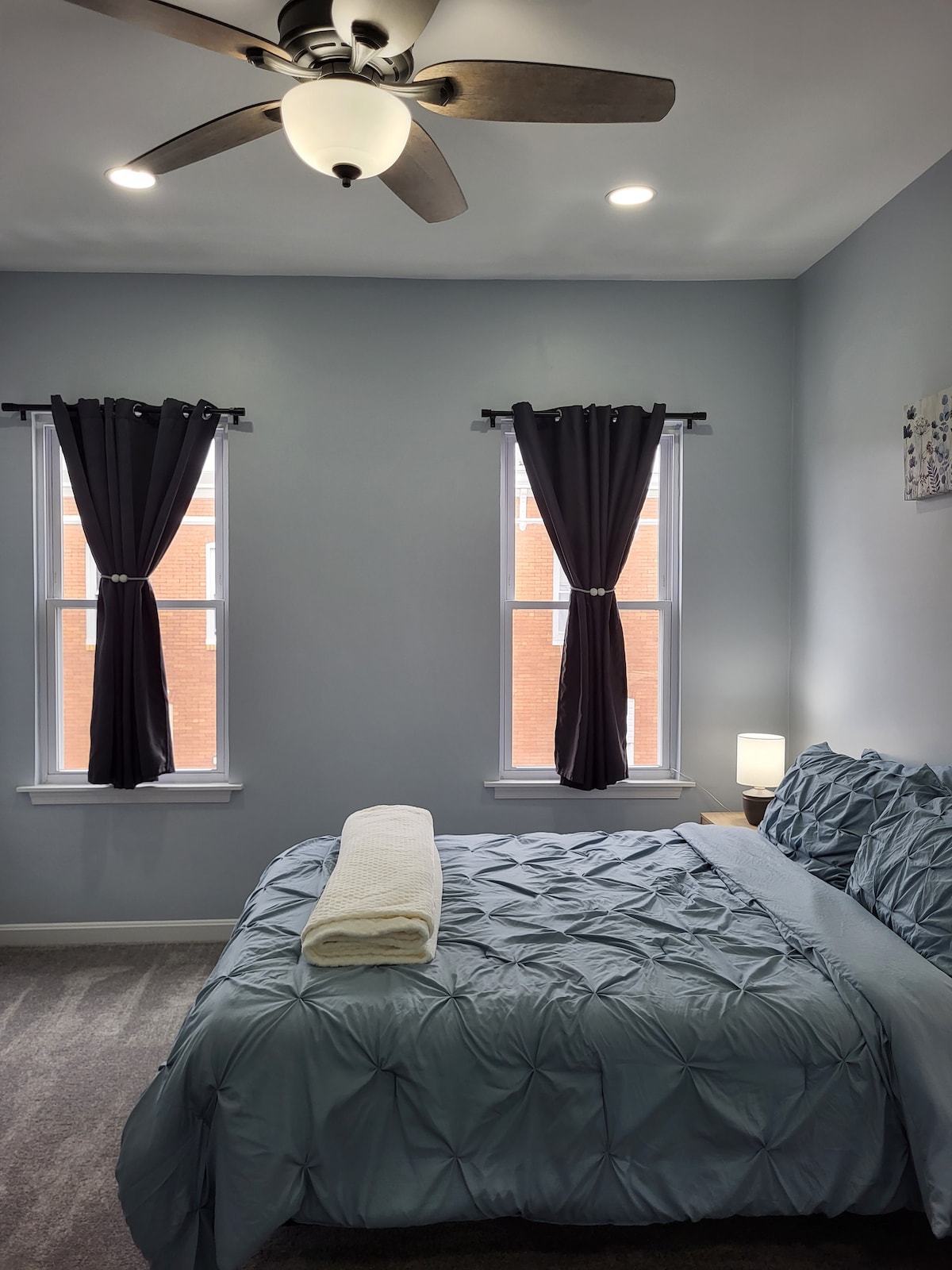 A well-lit master bedroom features a spacious bed with a textured blue bedspread. Two windows adorned with dark curtains allow natural light to filter in. A small bedside lamp sits next to the bed, and a ceiling fan is visible overhead.