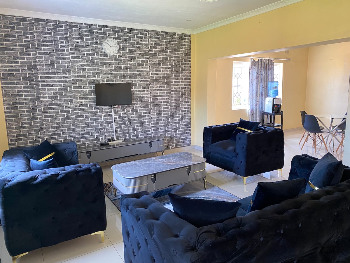 A modern living area is styled with upholstered seating in a deep color facing a low coffee table. A wall adorned with a textured design complements the space, while natural light filters through large windows covered by sheer curtains, enhancing the inviting atmosphere.