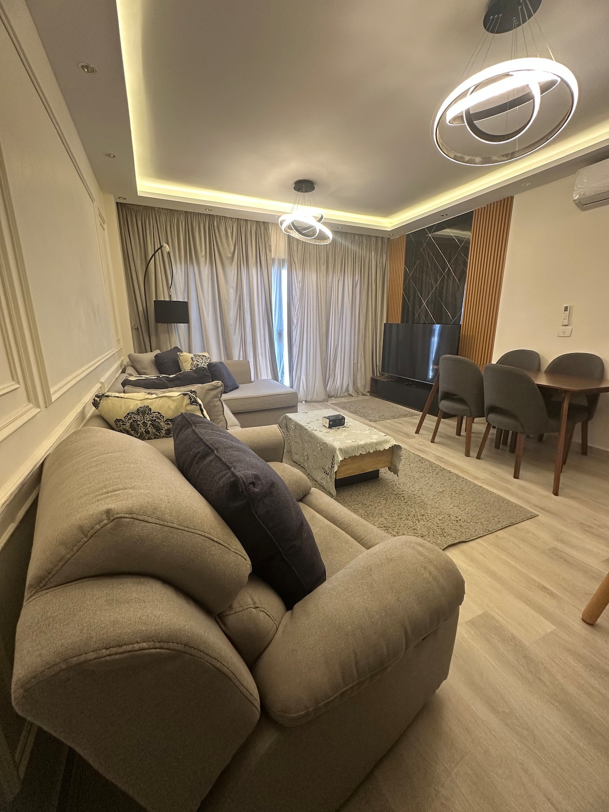 A modern living space is highlighted by neutral-toned sofas and stylish lighting fixtures. A dining area with a round table is visible, accompanied by upholstered chairs. Large, sheer curtains diffuse natural light from a window, while a television is placed on a media console.