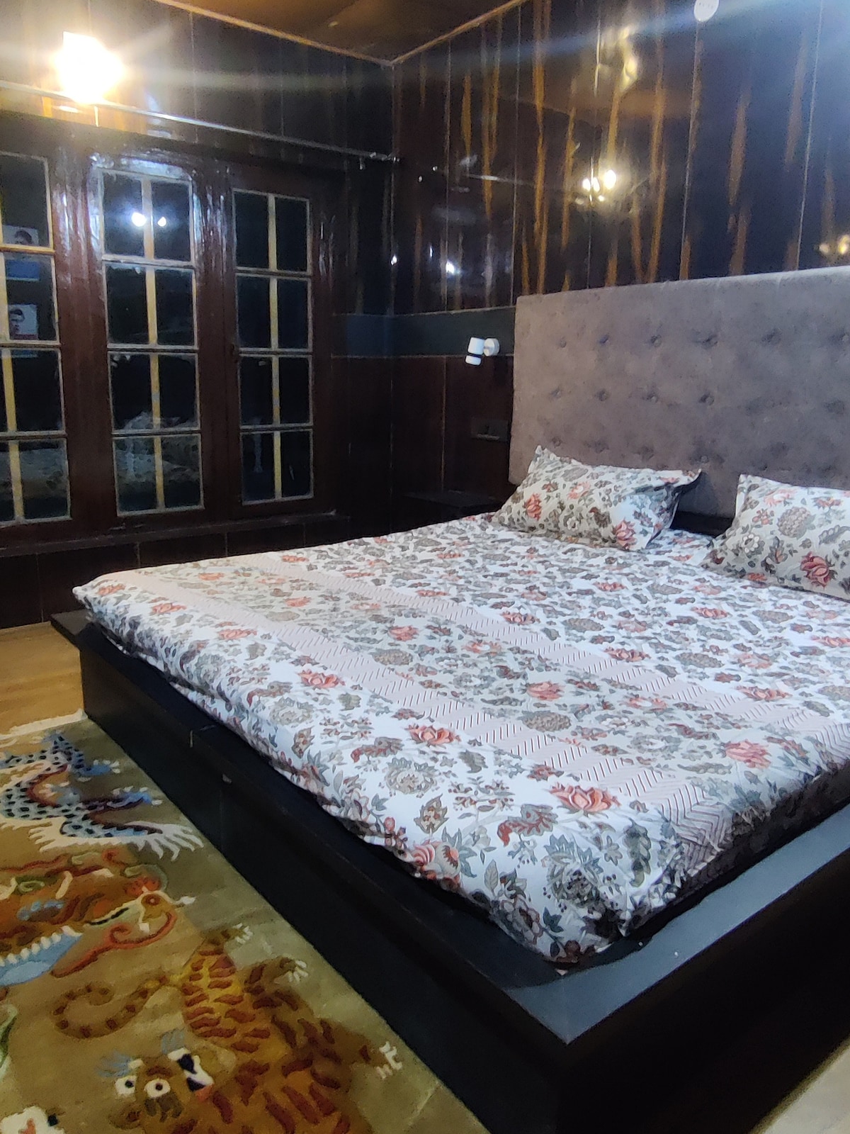 A spacious sleeping area features a large bed covered with floral-patterned bedding. The room's wooden paneling complements the dark floor, and multiple windows allow natural light to fill the space. A colorful area rug adds warmth to the room.