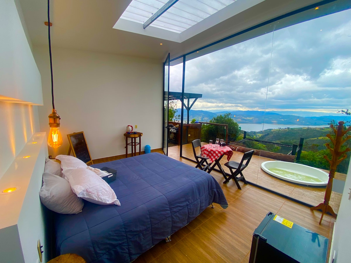 A stylish bedroom features a spacious bed with soft linens, complemented by a cozy seating area. Large glass doors open to a deck with a jacuzzi and views of the lake, while warm light is provided by a hanging pendant lamp.