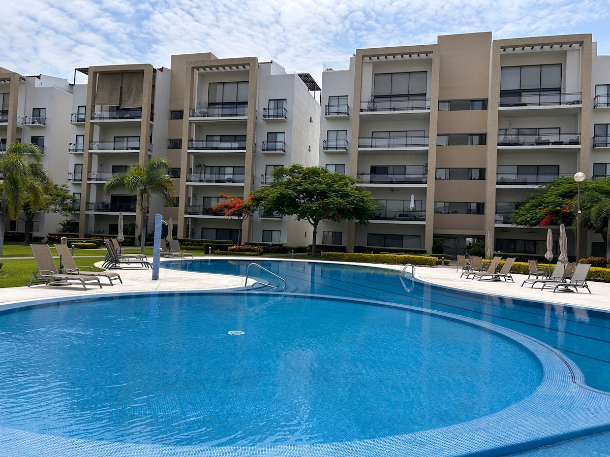 A spacious outdoor pool is surrounded by lounge chairs and neatly manicured lawns. The pool's circular design features gentle curves, while the adjacent multi-story buildings offer balconies with views of the area. Lush trees and flowering plants enhance the tranquil setting.