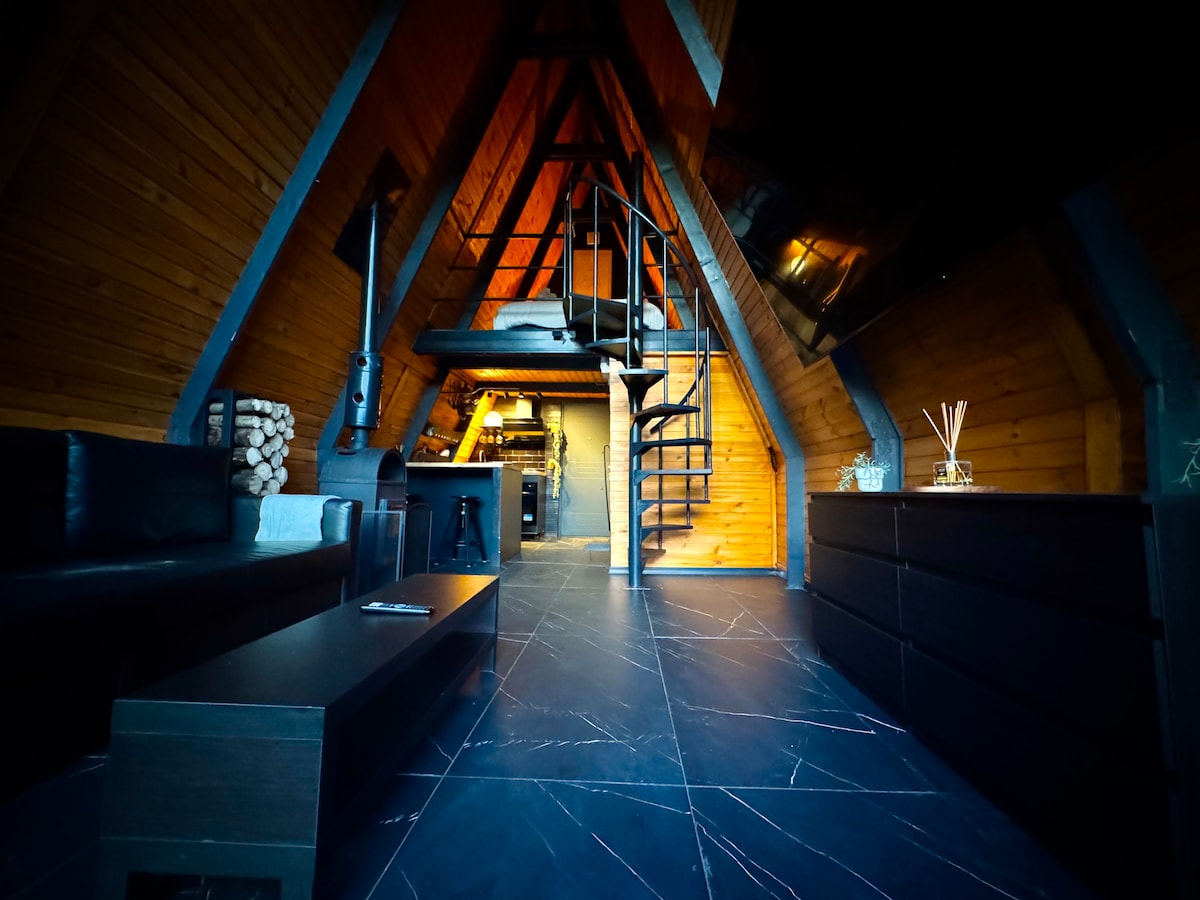 The open space features a spiral staircase leading to the upper level. Wooden walls create a cozy ambiance, complemented by dark tile flooring. A comfortable seating area is arranged around a coffee table, while a modern kitchen area is visible in the background, enhancing functionality.