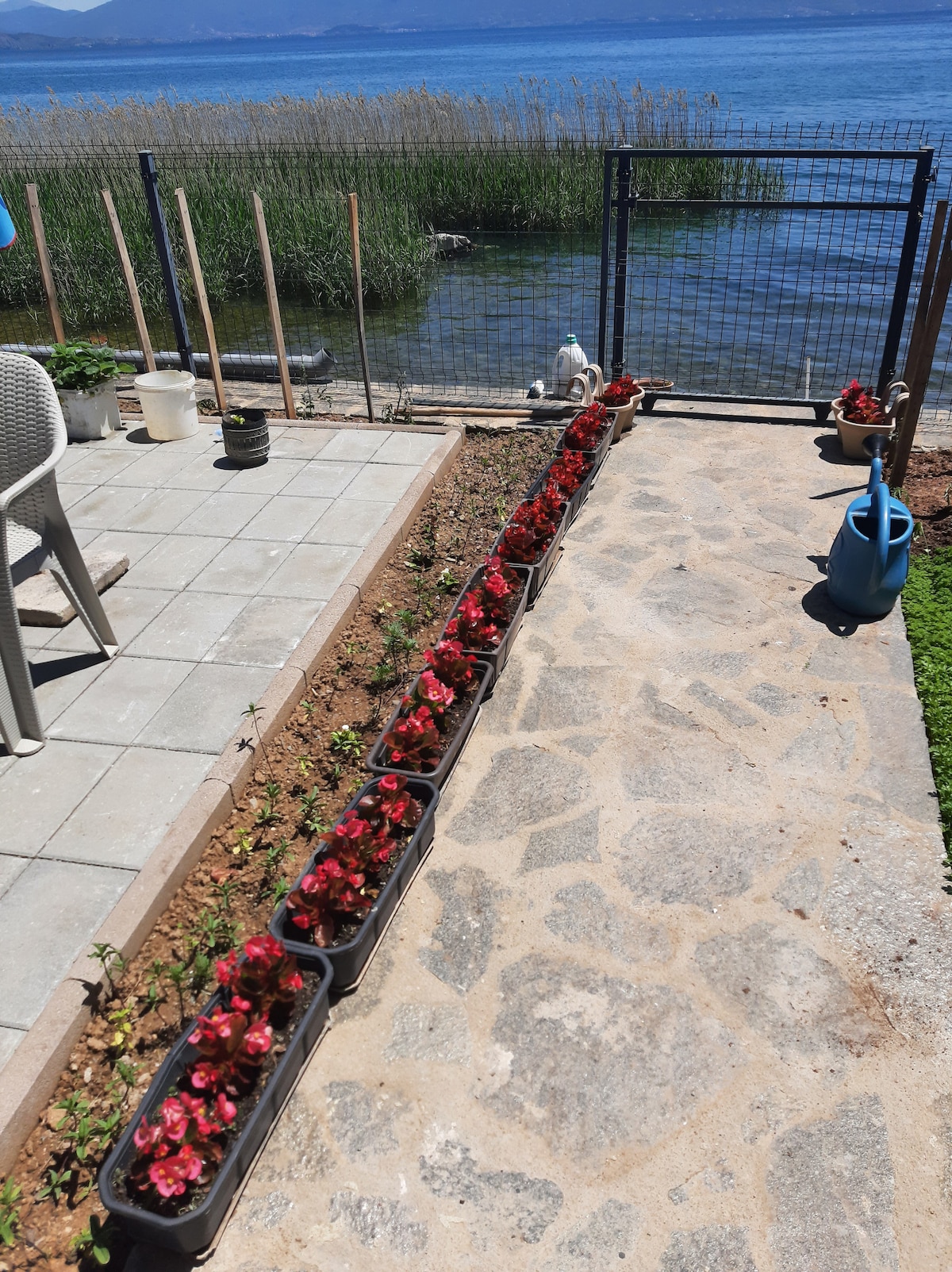 A pathway lined with flower boxes displays vibrant red blooms. Paved stones lead to a grassy area and a fence that overlooks a body of water. Nearby, a table and chairs are positioned on a patio, creating a welcoming outdoor space.