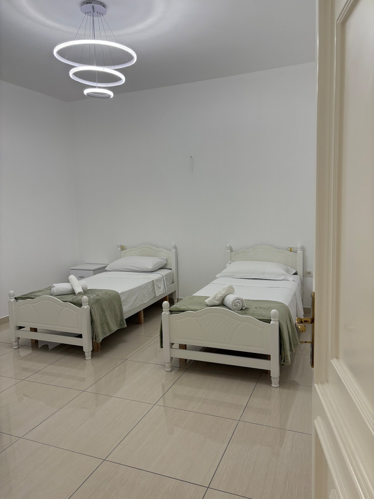 A well-lit room is furnished with three single beds, each made with clean white linens and light green throws. Two rolled towels are neatly placed on the beds. The walls are painted in a neutral tone, and the floor features glossy tiles that enhance the room's brightness.