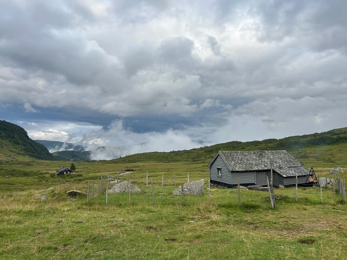 The image shows a traditional wooden cabin with a stone roof, set against a backdrop of rolling hills and a cloudy sky. Green meadows surround the cabin, with a wooden fence that adds to the rustic charm. Other distant structures are visible in the landscape.