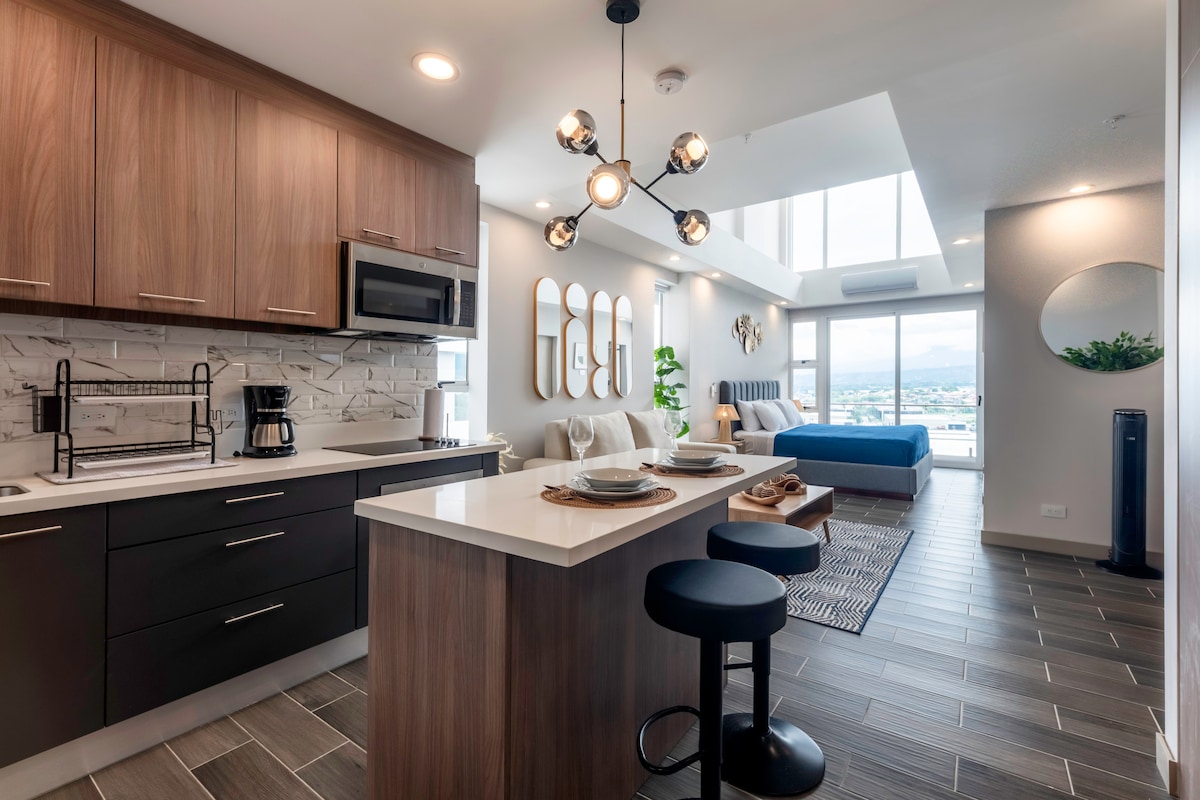 A modern studio apartment features a kitchen with wooden cabinetry and a countertop island with seating. Natural light floods the space through large windows, illuminating the living and sleeping areas, which include a cozy bed and stylish decor. A decorative plant enhances the ambiance.
