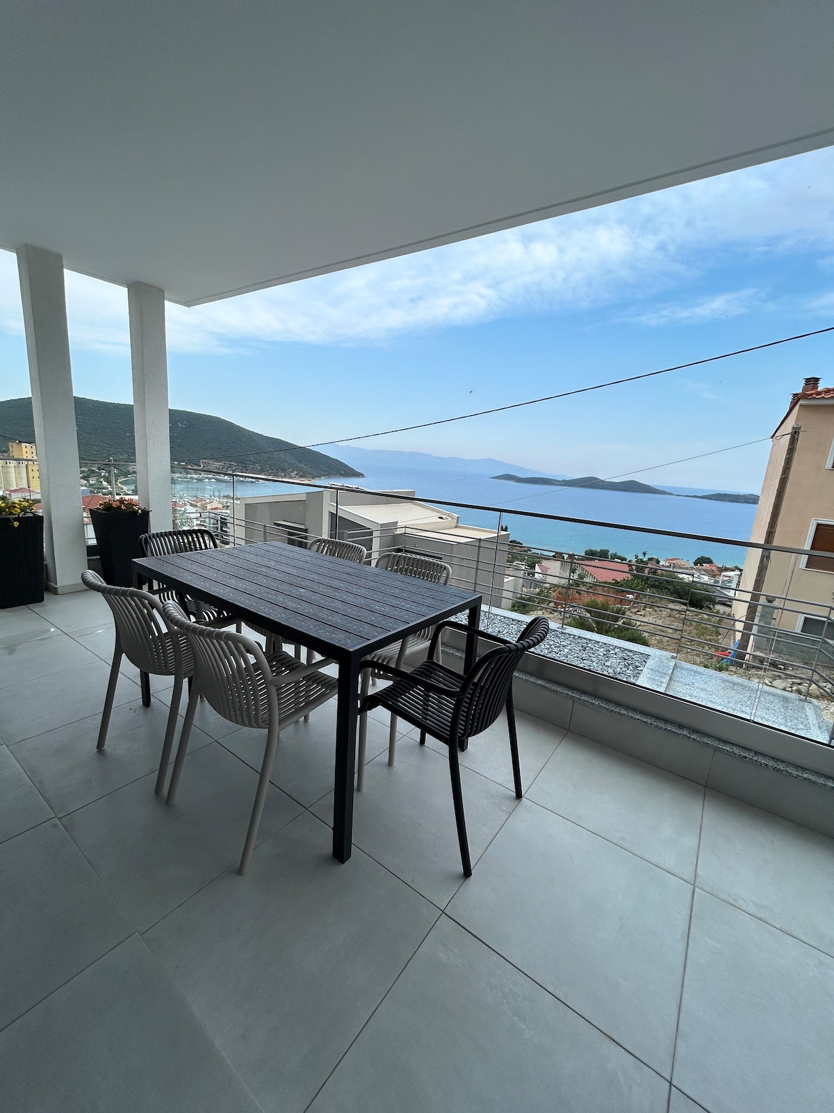 An outdoor terrace is furnished with a rectangular dining table surrounded by six chairs, offering a scenic view of the sea and mountains in the distance. Large windows allow natural light into the space, which is designed with clean, modern lines.