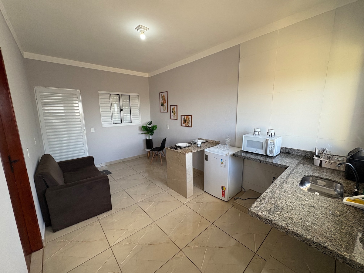 A spacious kitchenette is seen, featuring a granite countertop, a microwave, and a compact refrigerator. A small dining table with black chairs is positioned next to the kitchenette. A dark sofa is placed in the corner, complemented by light tile flooring and muted wall colors.