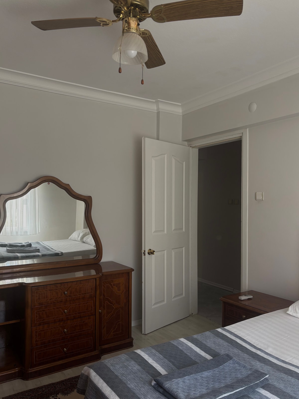 A cozy bedroom is equipped with a spacious bed, complemented by a decorative gray and white blanket. A vintage wooden dresser with a mirror is positioned against the wall, reflecting natural light from a nearby window. A door leads to another room.