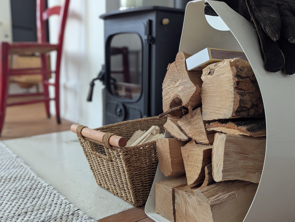 A woven basket filled with neatly arranged firewood sits beside a black wood-burning stove. The stove features a rounded design and a cozy ambiance. A red chair is positioned in the background, contributing to the warm environment of the flat.
