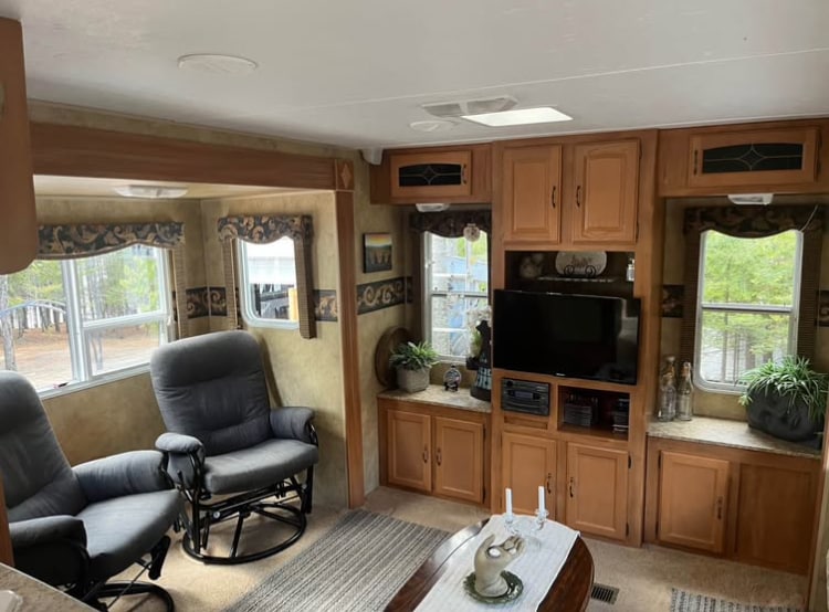 The living area features two comfortable rocking chairs positioned near a media center with a television. Natural light filters in through multiple windows adorned with decorative valances, while potted plants add greenery to the space. A neutral-toned area rug anchors the room.