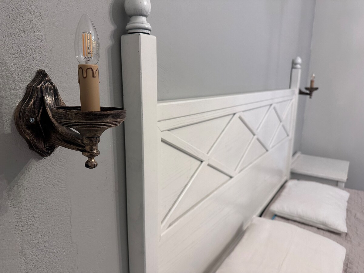 Stylish wall-mounted sconces are positioned on either side of a headboard crafted from white wood, featuring a geometric design. Soft pillows are arranged on the bed, and light from the sconces casts a gentle glow on the light gray wall.