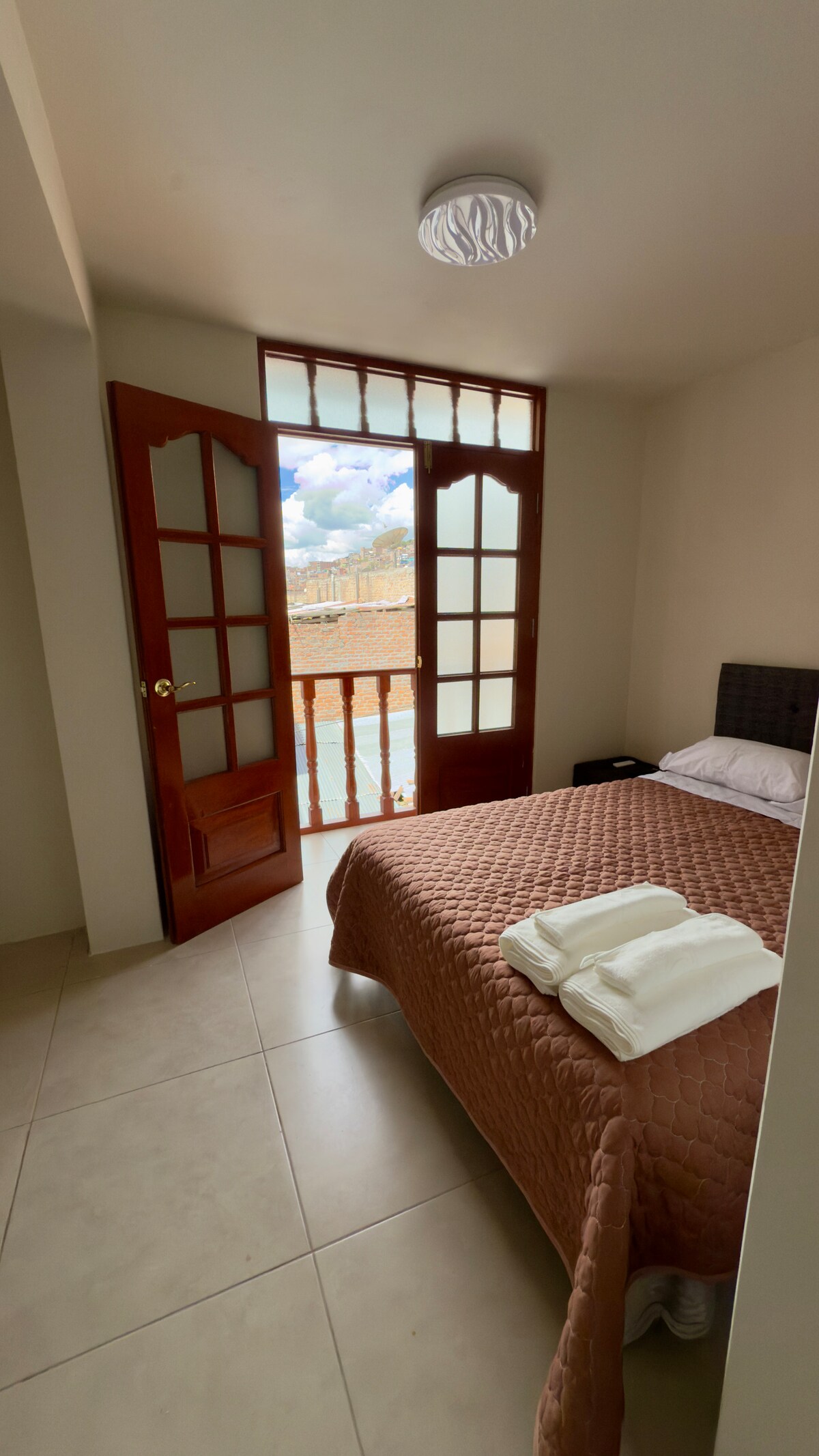 An inviting bedroom features a spacious bed with a patterned blanket. Large glass doors open to a balcony, allowing natural light to fill the room. Crisp white towels are neatly arranged on the bed, and a neutral-colored floor complements the decor.