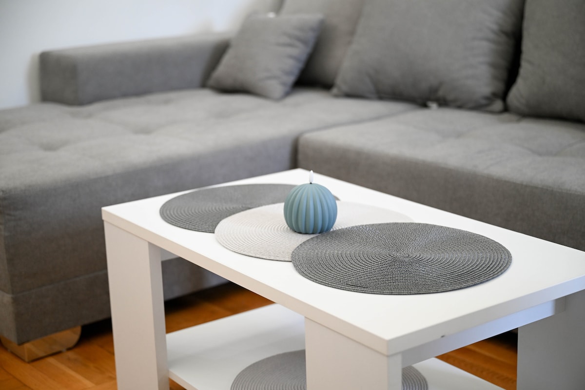A neutral-toned sofa is paired with a white coffee table. The table is adorned with three round placemats in various shades of gray and a decorative candle in a soft blue hue, creating a balanced and simple centerpiece.