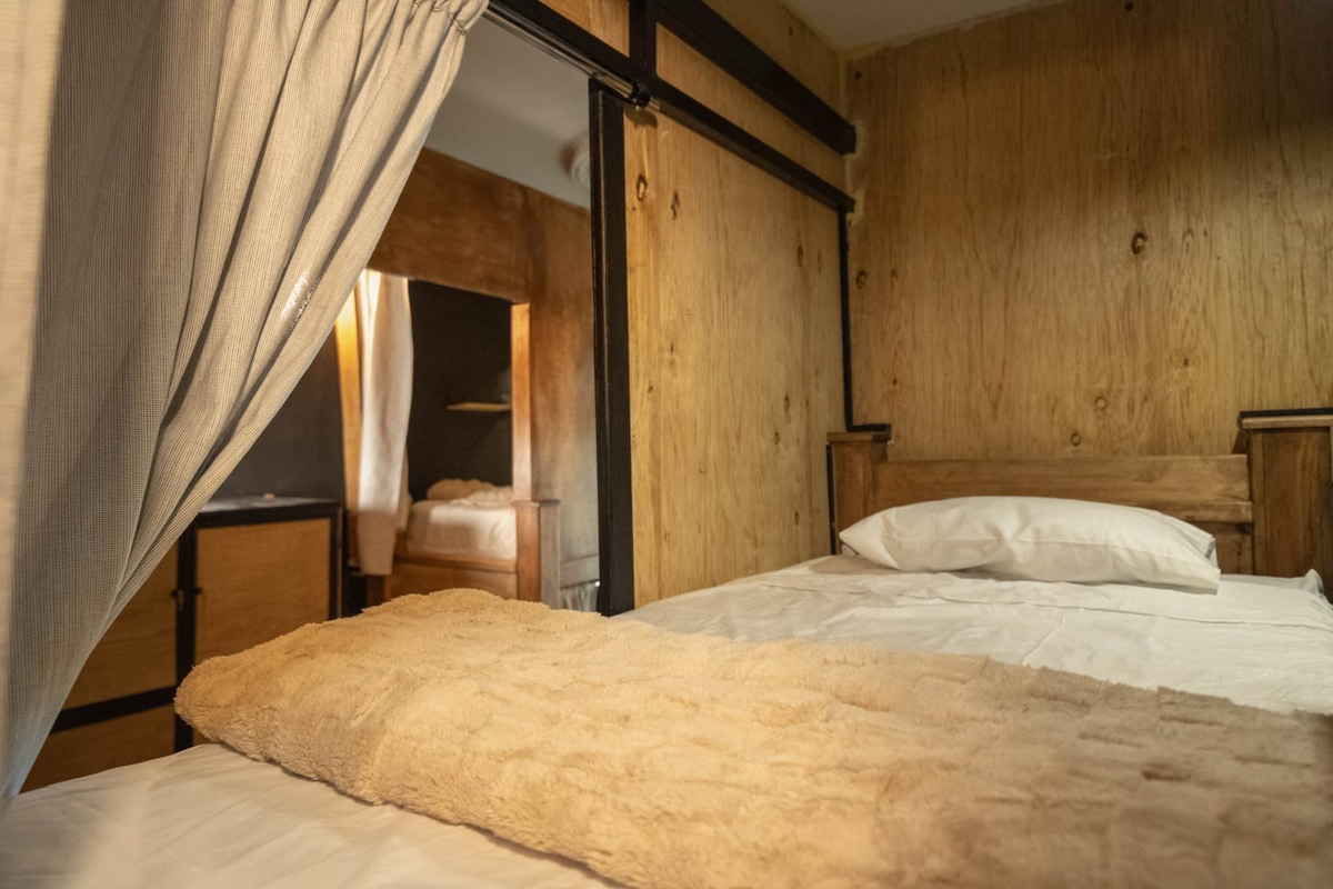 A modern capsule accommodation is shown, featuring a spacious bed with a soft blanket draped across it. The wooden walls provide a natural feel, while a curtain offers privacy. Additional sleeping areas can be glimpsed in the background, enhancing the cabin's functionality.