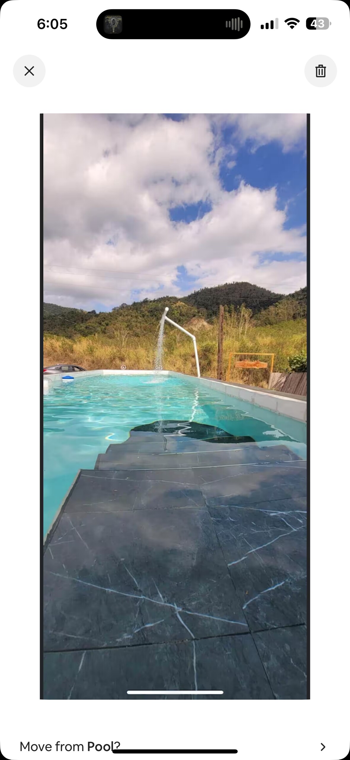 A swimming pool with clear turquoise water is shown, surrounded by natural greenery and rocks. A gentle waterfall feature is visible, cascading into the pool. The landscape in the background includes hills under a partly cloudy sky.