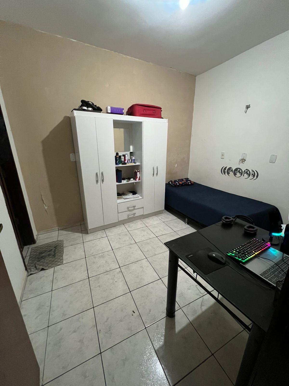 A single room is presented with light-colored walls and tiled flooring. A spacious bed with dark bedding occupies one corner. A white wardrobe displays personal items, while a black table with a computer and accessories is positioned near the room's entrance.