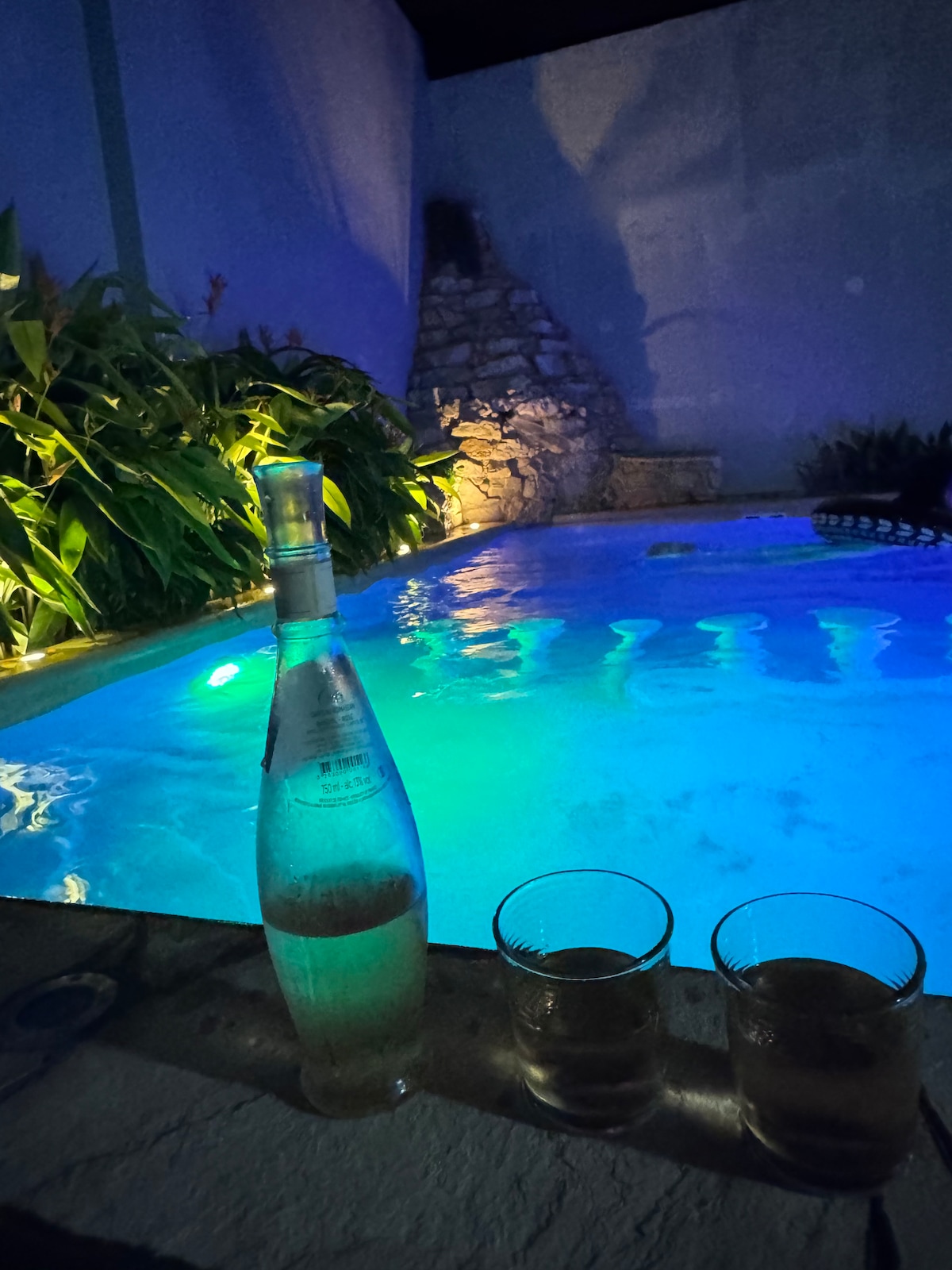 A serene pool area is softly illuminated in shades of blue, surrounded by lush greenery. A bottle of sparkling water is positioned next to two glasses filled with a beverage, suggesting a tranquil atmosphere for relaxation.