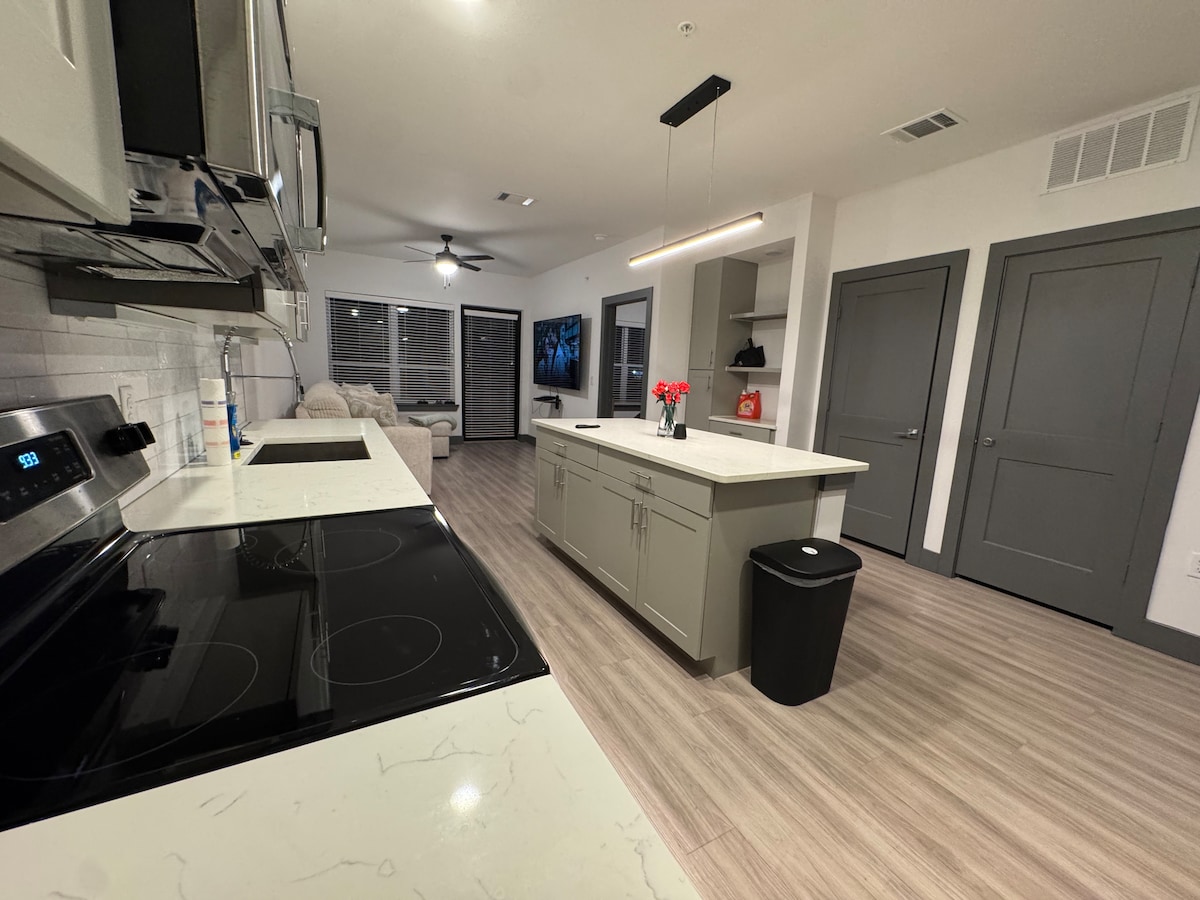 A modern kitchen is highlighted, featuring sleek gray cabinetry and a large island with a white countertop. Stainless steel appliances, including an oven and microwave, are visible. A black trash bin is positioned nearby, and a dining area is seen in the background through an open space.