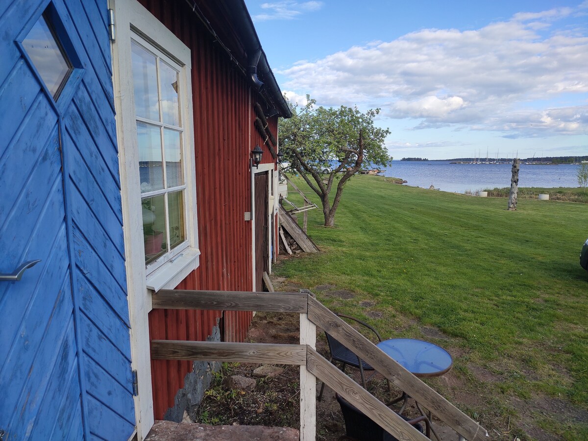 A rustic exterior showcases a vibrant blue door against a red wooden wall. A grassy lawn leads towards a serene waterfront with boats in the distance. A nearby tree provides shade and a sense of tranquility, enhancing the natural surroundings.