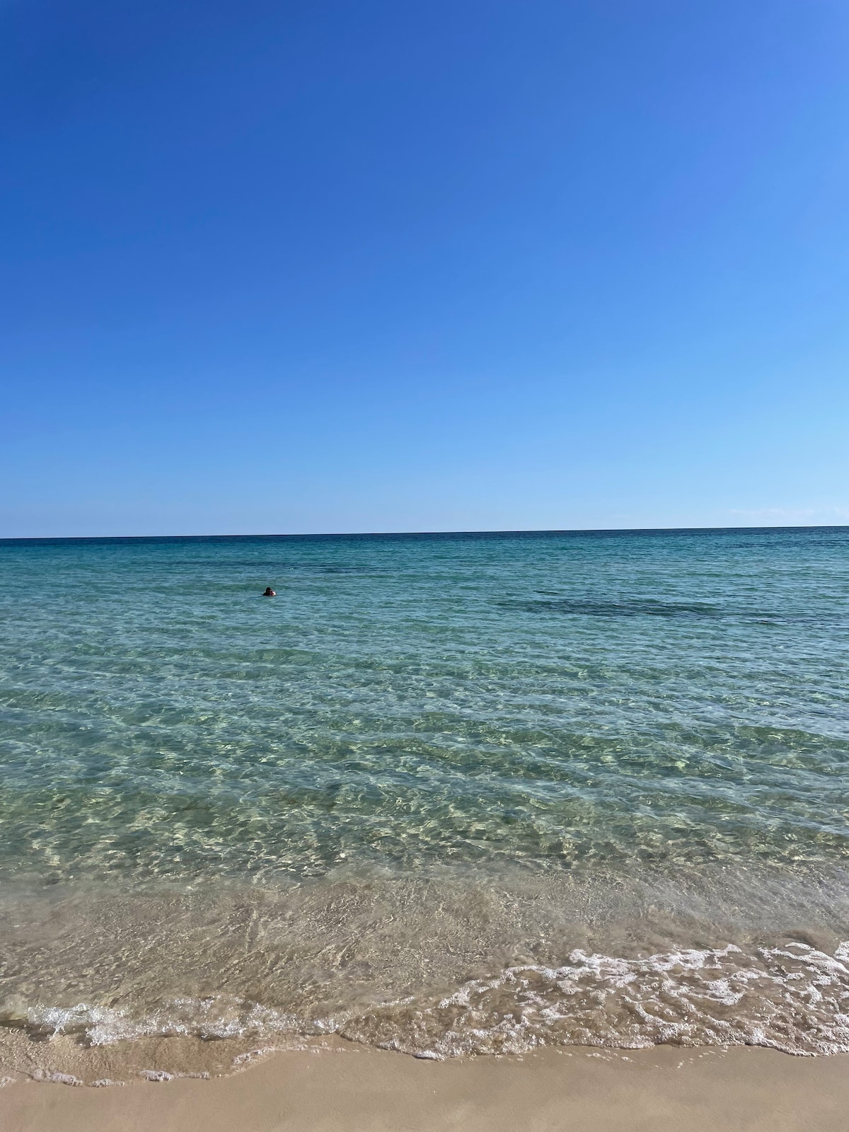 A serene beach scene features clear, shallow waters reflecting the blue sky. Gentle waves lap against the soft, sandy shoreline, inviting relaxation. A small figure can be seen swimming in the distance, illustrating the tranquil atmosphere of this coastal destination.