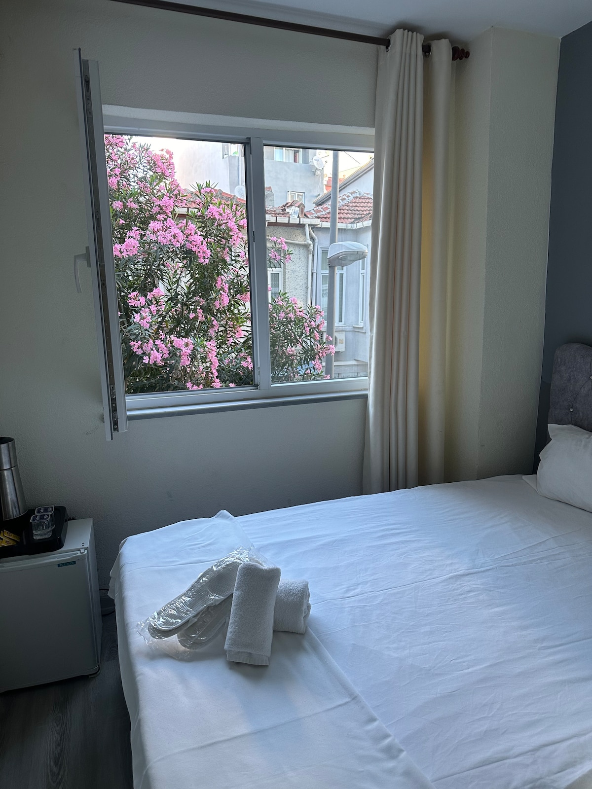 A bright sleeping area features a spacious bed with white linens. A window offers a view of flowering pink bougainvillea outside, while sheer curtains allow soft daylight to filter in. A small fridge is positioned next to the bedside, contributing to the room's convenience.