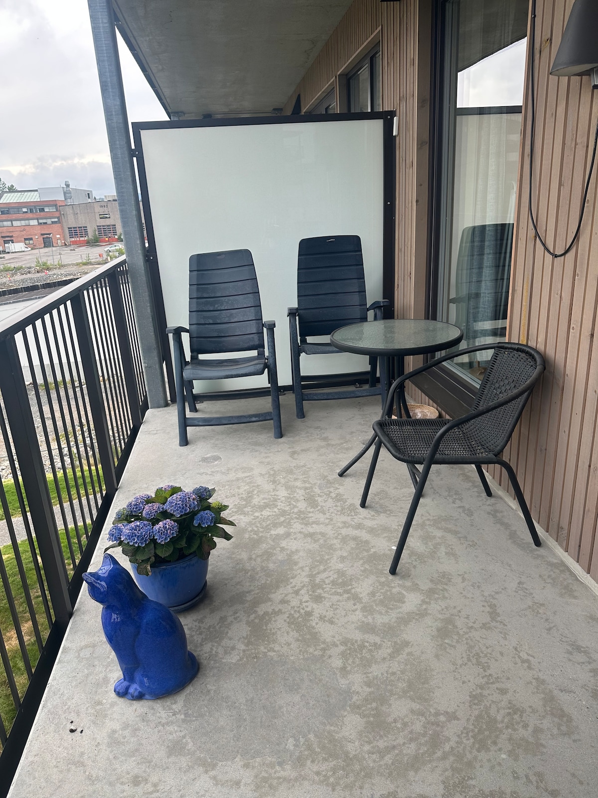 A cozy balcony area features two black rocking chairs and a glass-topped table. A rattan chair and a blue ceramic planter with blooming flowers are also present. The balcony is enclosed by a glass partition, providing a sense of privacy while allowing views of the surroundings.